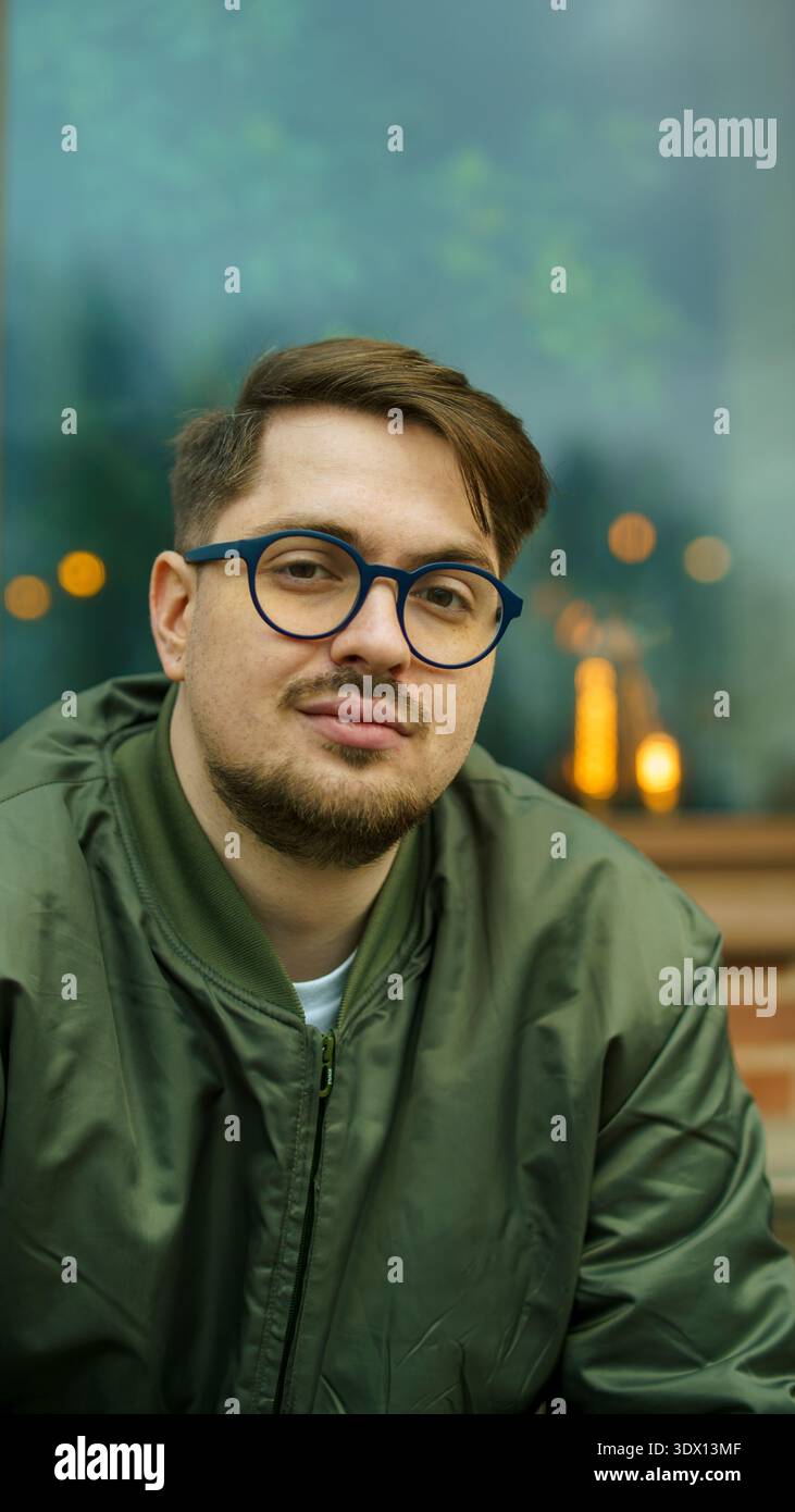 Ritratto verticale di un affascinante hipster caucasico barbuto in occhiali da vista e una giacca verde che posa all'aperto davanti a una finestra scura. Foto Stock