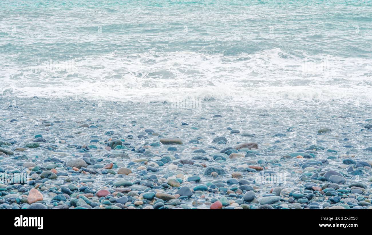 Ciottoli lisci e colorati sparsi su una spiaggia con onde delicate che si lavano sopra di loro Foto Stock