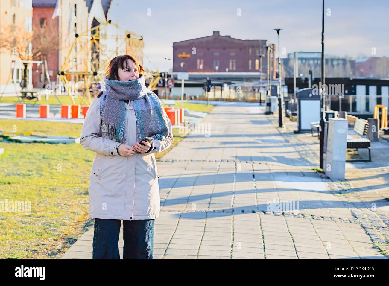 La figura femminile con un caldo cappotto e una sciarpa si erge su un sentiero soleggiato, tenendo in mano lo smartphone, con panche ed erba sullo sfondo, per catturare un po' di relax Foto Stock