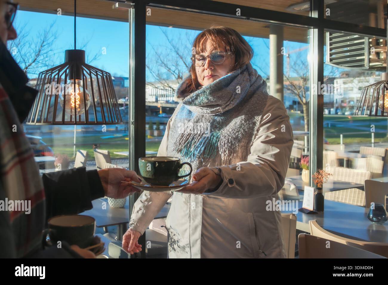 Una cliente donna in una caffetteria, vestita con calore, sta consegnando una tazza di caffè, creando un'interazione amichevole in un ambiente luminoso e invitante Foto Stock