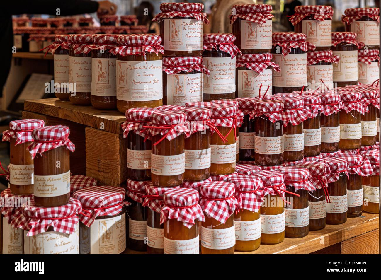 Barcellona, Spagna - 24 gennaio 2026: Barattoli impilati di marmellata artigianale con tradizionali rivestimenti in tessuto rosso a scacchi su un banco di legno. Foto Stock