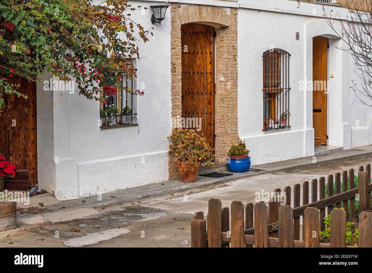 Tranquilla strada residenziale con facciata in stucco bianco, porte in legno, finestre a chiocciola, piante in vaso, vite con fiori rossi e una piccola recinzione in legno i Foto Stock