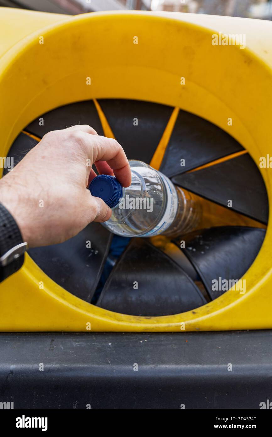 Mano che tiene una bottiglia di plastica sopra l'apertura di un contenitore di riciclaggio giallo, pronta a lasciarla cadere Foto Stock