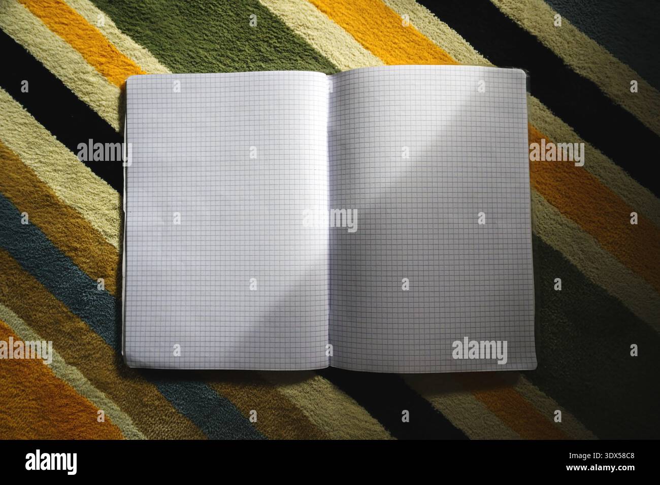 Notebook quadrato vuoto aperto posizionato su un tappeto colorato a righe con luce naturale e ombre soffuse. Composizione minima ideale per la scrittura, la pianificazione, Foto Stock