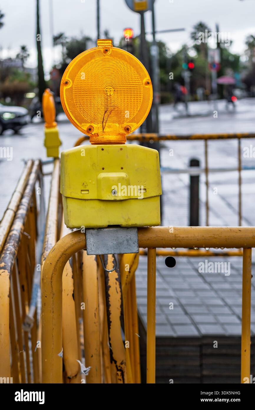 Primo piano della barriera gialla con spia ambra su una strada urbana piovosa, marciapiedi bagnati e semafori sfocati sullo sfondo. Foto Stock