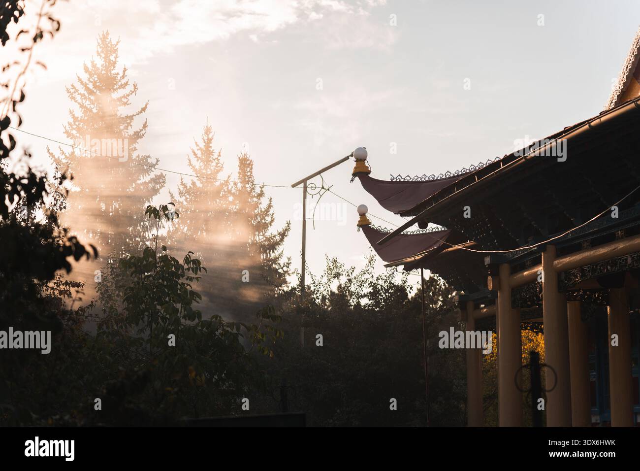 I raggi del sole dorato filtrano delicatamente attraverso gli alberi accanto a un tradizionale tempio buddista in Kirghizistan. I dettagli decorati del tetto creano una spiritua tranquilla Foto Stock