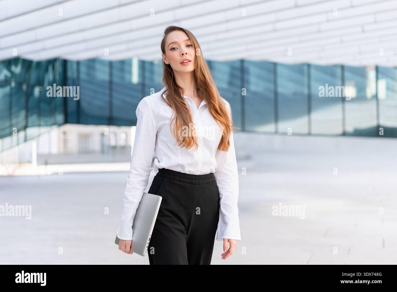 Donna d'affari sicura con camicia bianca e pantaloni neri in piedi con un computer portatile chiuso vicino a un moderno edificio per uffici. Fiducia professionale e concetto di lavoro aziendale Foto Stock