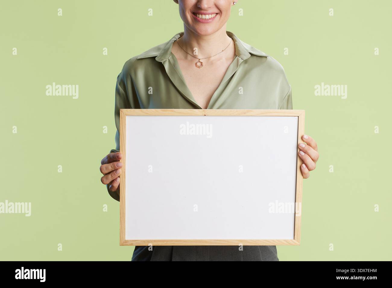 Donna sorridente che tiene in mano una lavagna bianca, indossa una blusa verde oliva e una collana. Atmosfera positiva e invitante. Foto Stock