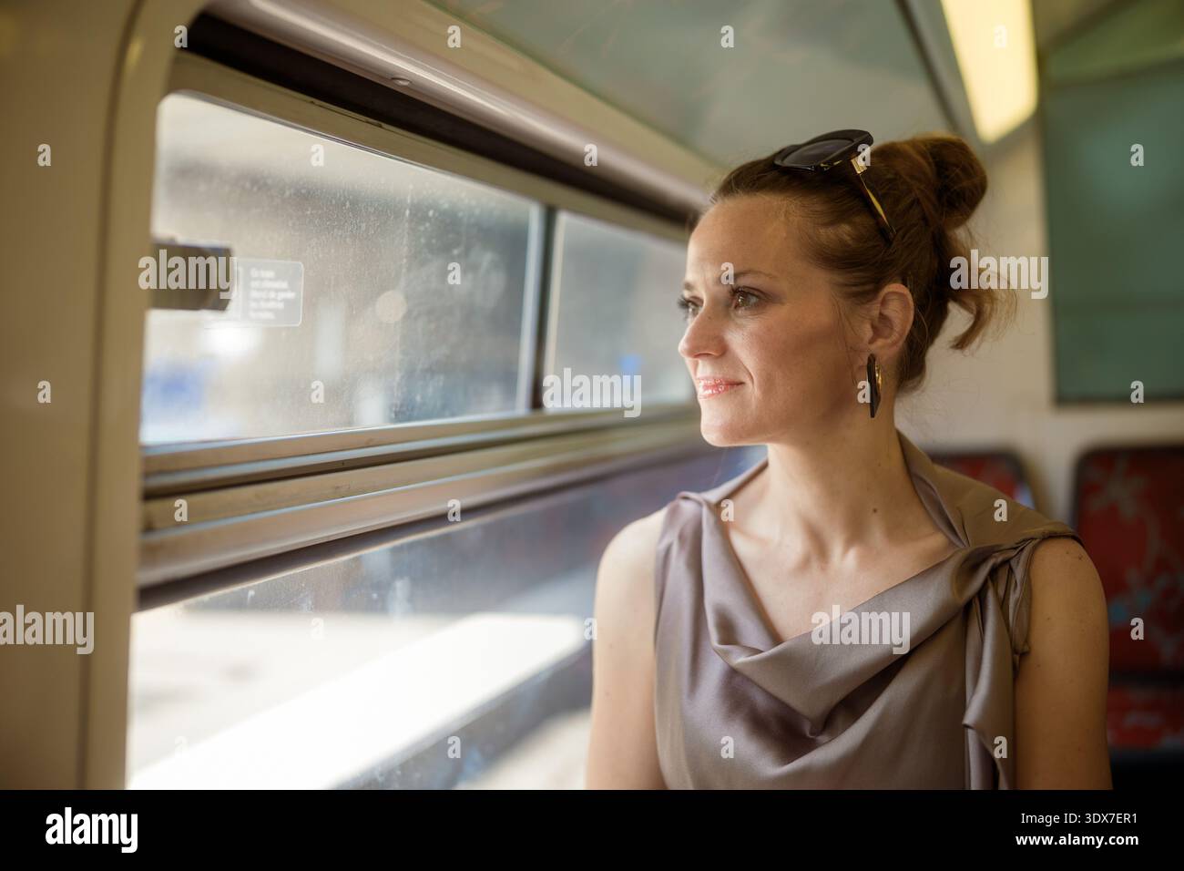 Una donna elegante in un abito grigio in raso guarda con attenzione fuori dal finestrino del treno, abbracciando i momenti contemplativi del suo viaggio da solo. Il suo demo avvelenato Foto Stock