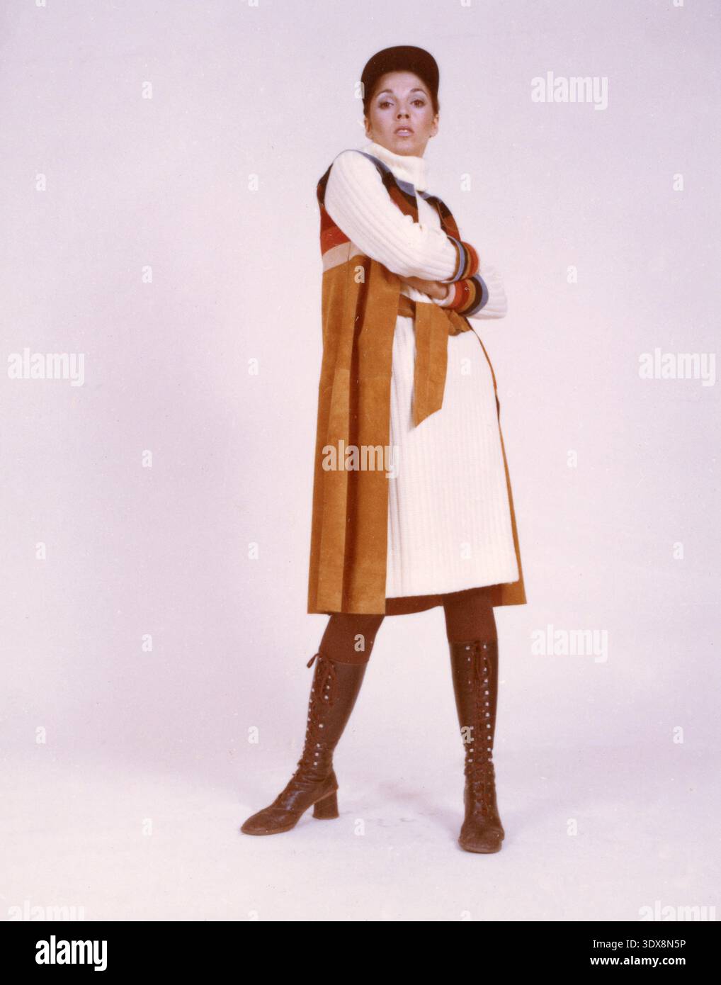 Modello che indossa un abito in maglione dolcevita a coste con un gilet longuette in pelle scamosciata color abbronzatura con pannelli a righe tonalità terra, un berretto piatto coordinato e stivali con lacci marroni, Italia anni '1970 Foto Stock