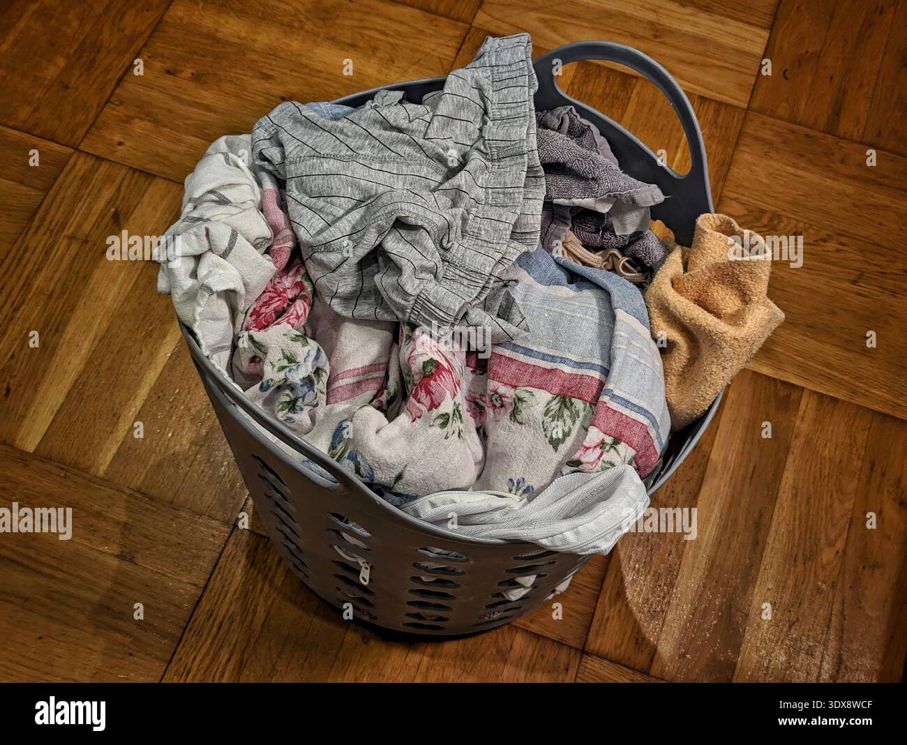 Cestino per la lavanderia traboccante sul pavimento parquet, versamenti di tessuti misti e asciugamani, maglione grigio drappeggiato sul bordo, bordi in fogli floreali visibili, caldo interno Foto Stock