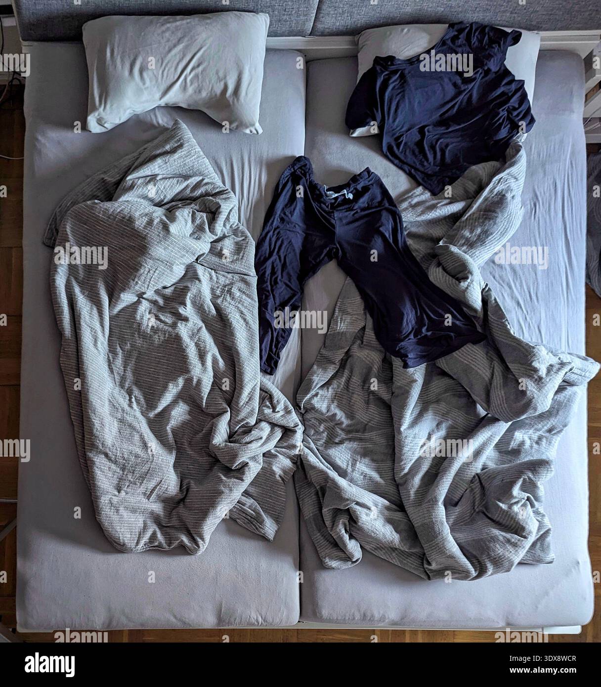 Letto matrimoniale non realizzato con lenzuola sgualcite, due camicie navy spalmate al centro, piumino beige intrecciato, cuscini spinti da parte, luce soffusa sopra la testa, intima Foto Stock