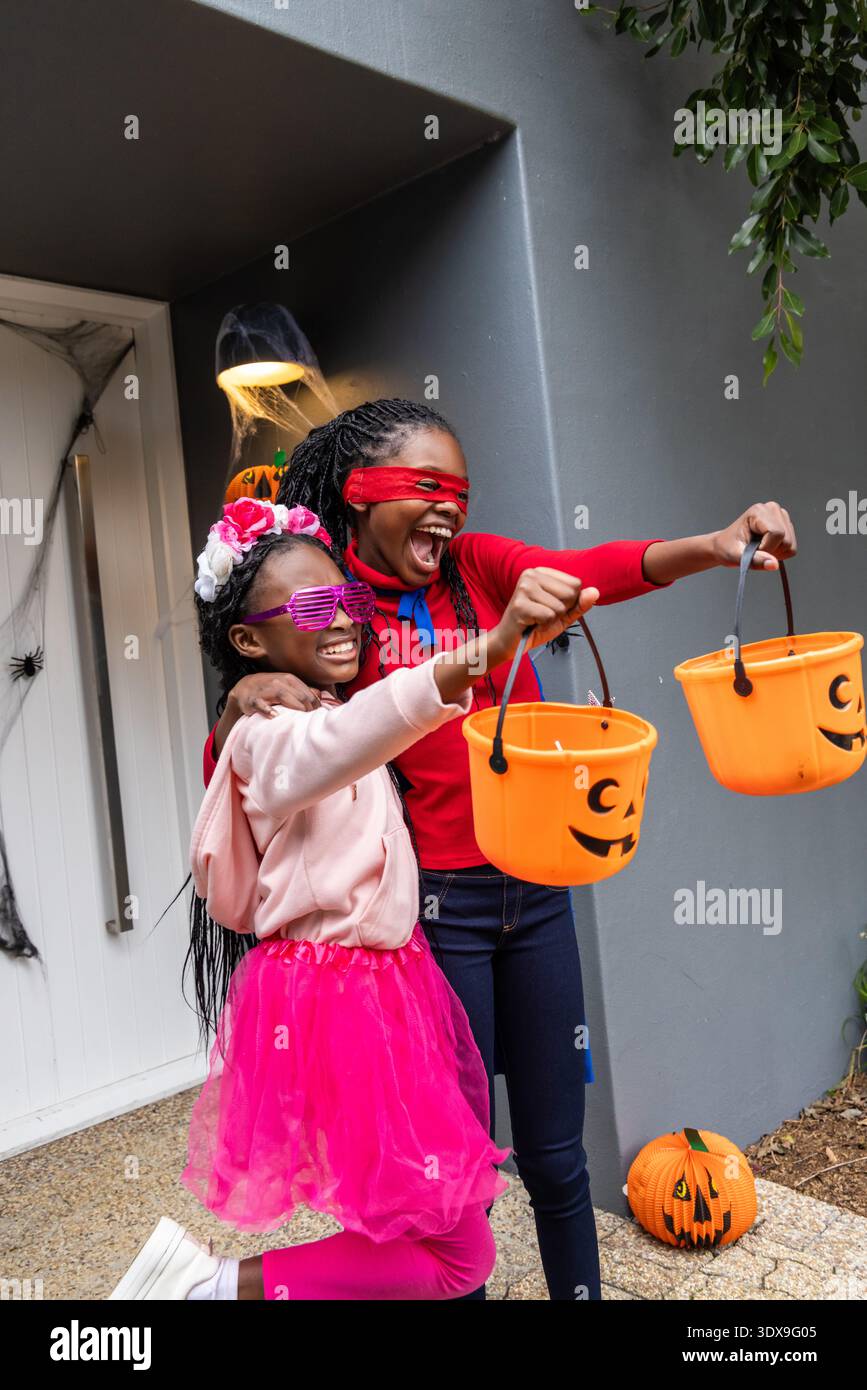 Bambina afroamericana e adolescente vestita con costumi, tenendo secchi di zucca arancioni sul portico Foto Stock