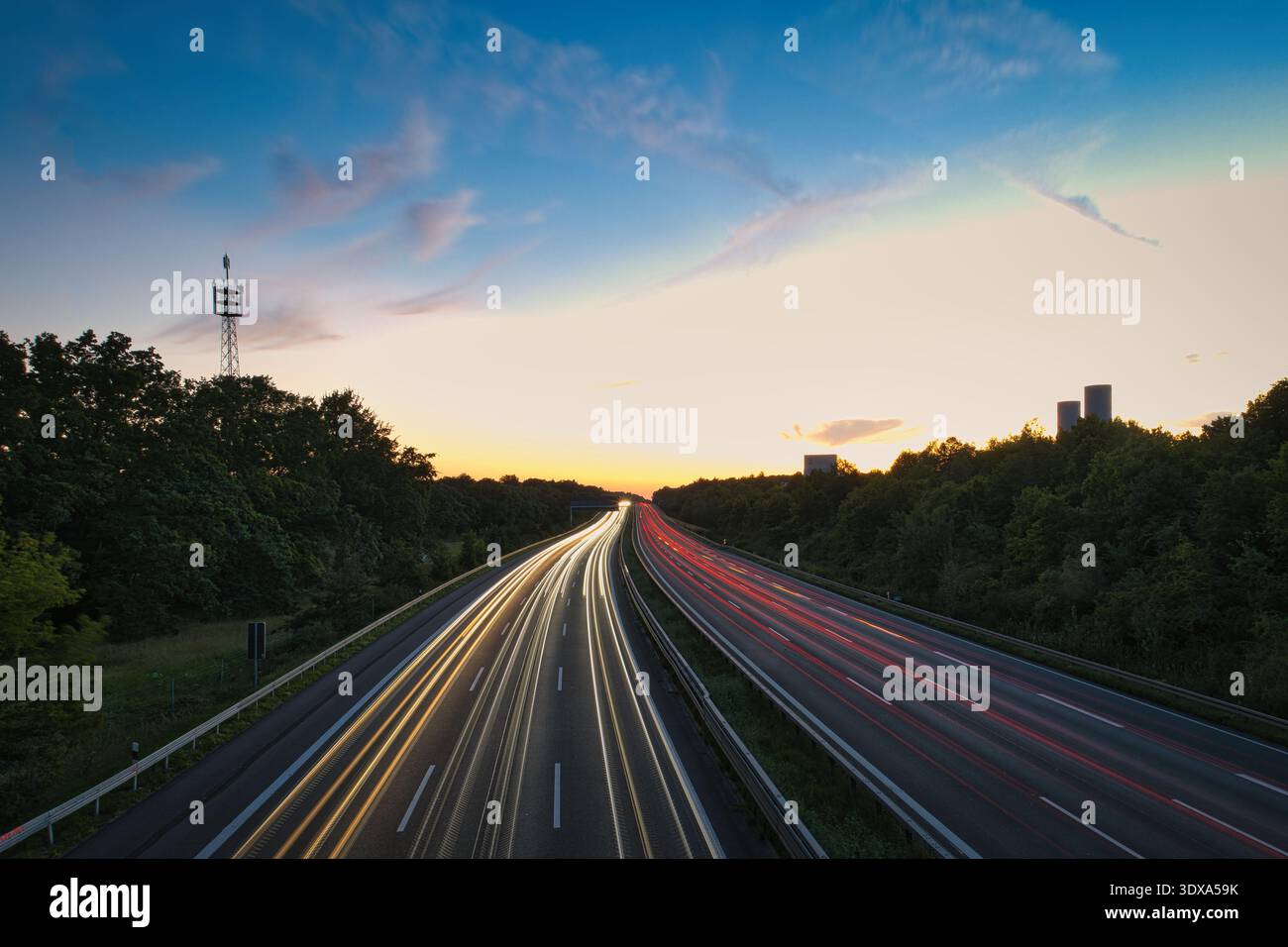 Lunga esposizione del traffico autostradale al tramonto, con percorsi leggeri dai fari e dalle luci posteriori dell'auto contro il cielo del tramonto e le sagome degli alberi Foto Stock