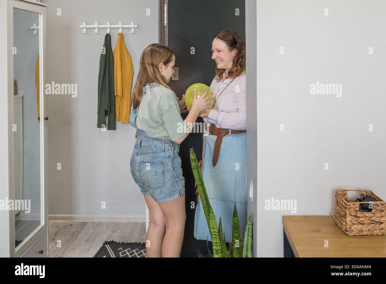 Mamma e figlia stanno all'ingresso di un appartamento moderno. Un amico sta tenendo in mano un frutto grande mentre sorride e sembra che si goda un conver amichevole Foto Stock
