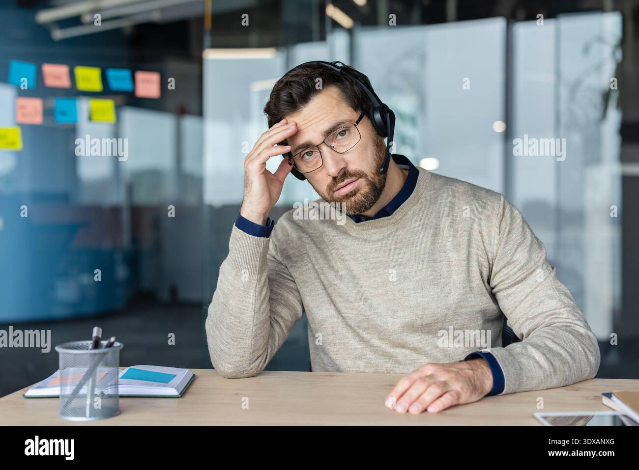 Frustrato da un operatore di contact centre che indossa cuffie e occhiali, tiene la testa, prova stress e mal di testa mentre guarda la fotocamera in un ufficio moderno Foto Stock
