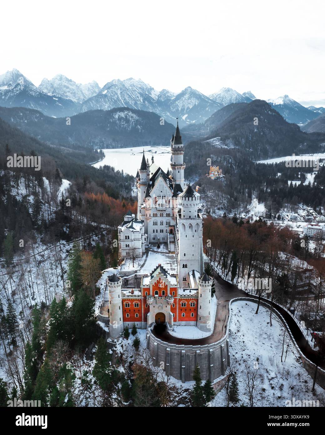 Vista aerea delle guglie del castello di Neuschwanstein che perforano il cielo tra foreste innevate e vette smerigliate lontane, una visione fiabesca incisa nel vento Foto Stock