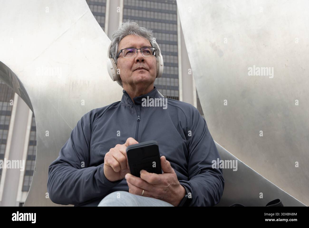 Uomo asiatico anziano seduto su una scultura di metallo in plaza, che usa lo smartphone con le cuffie bianche Foto Stock