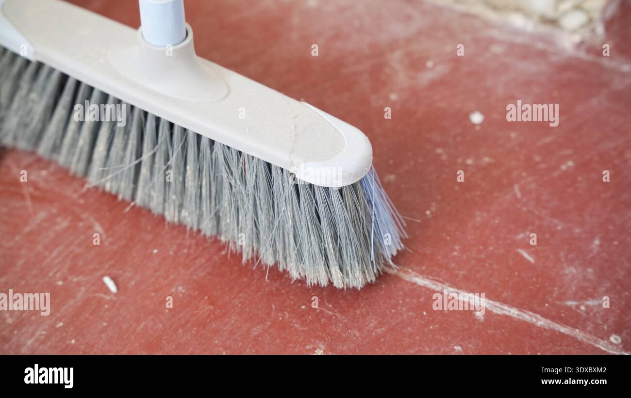 Scopa per uso domestico con setole grigie che spazzano la superficie del pavimento rosso polveroso durante la pulizia dell'appartamento. Foto Stock