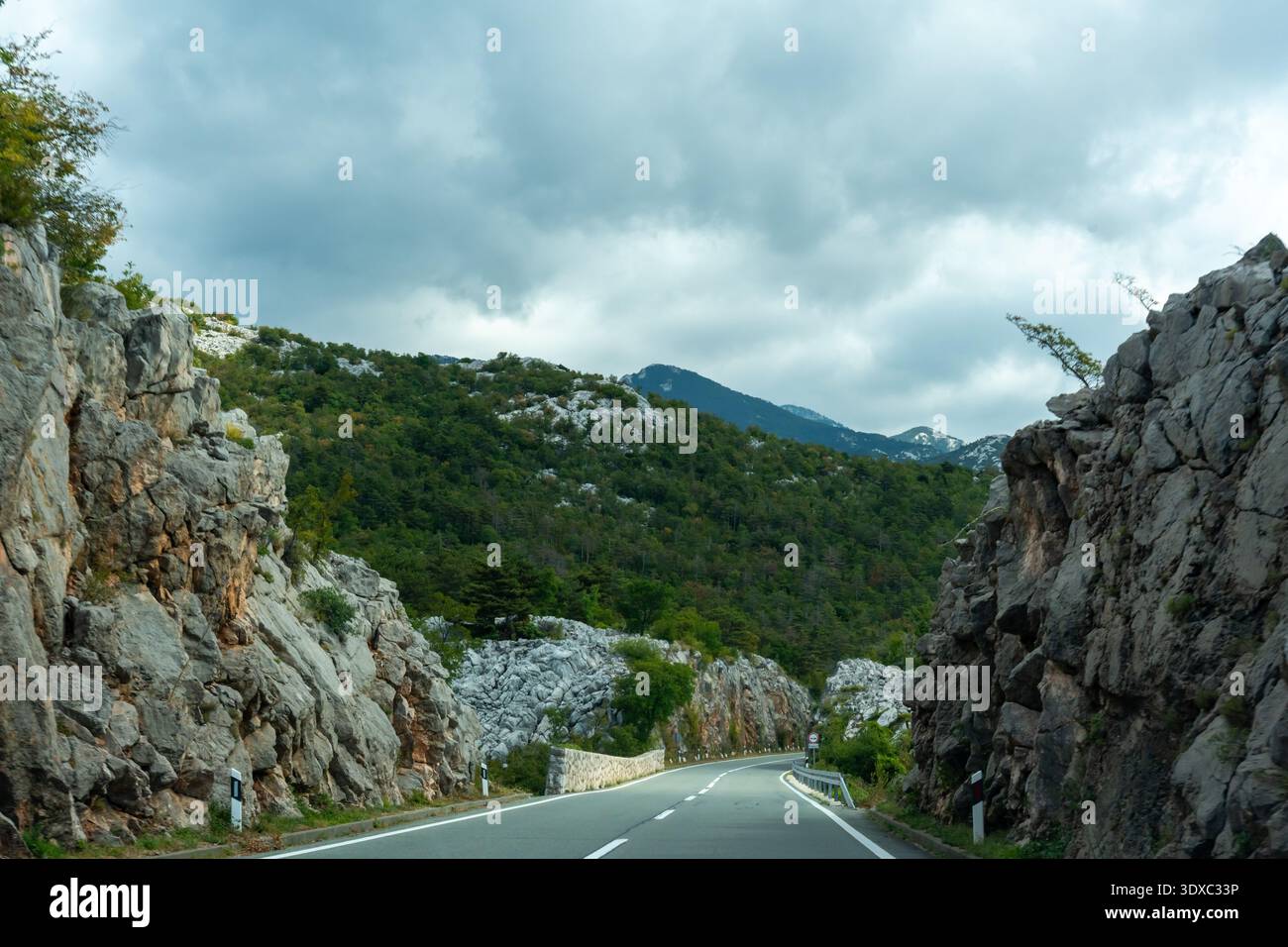 Autostrada di montagna vuota tra due massicce pareti di roccia grigia con lussureggianti pendii boscosi e un suggestivo cielo tempestoso davanti a te. Concetto di guida avventurosa, selvaggia Foto Stock