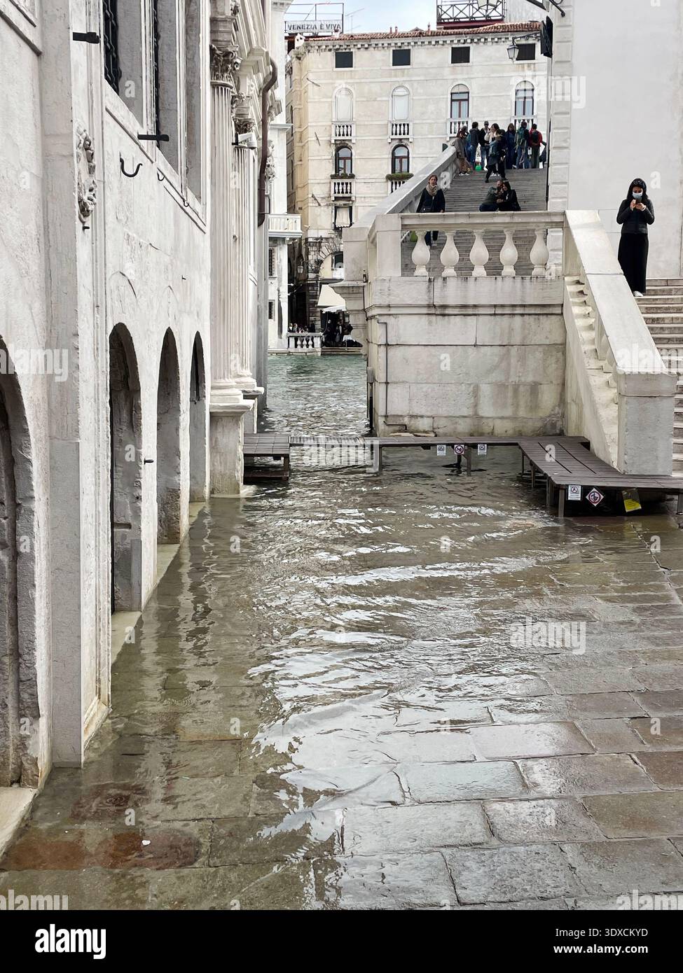 L'acqua alta inonda un passaggio pedonale in pietra vicino a un canale mentre i pedoni attraversano un piccolo ponte durante l'acqua alta a Venezia. - Immagine stock catturata con smartphone
