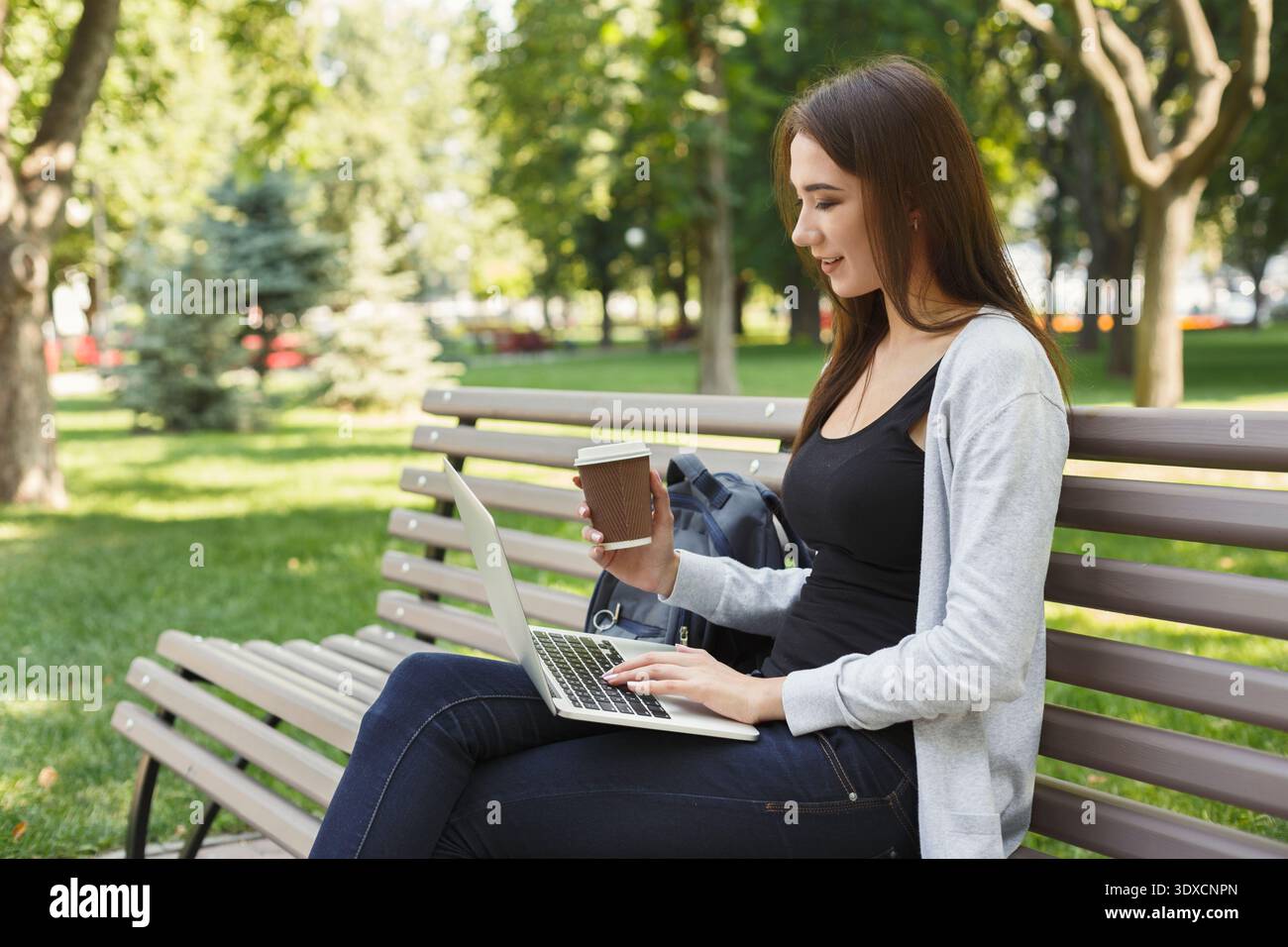 Una giovane donna lavora su un notebook mentre è seduta su una panchina in un parco con il caffè in mano Foto Stock