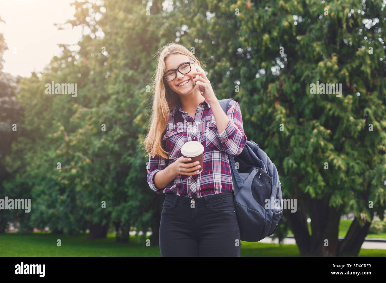Cheerful Girl parla al telefono mentre tiene il caffè in Park circondato da alberi Foto Stock