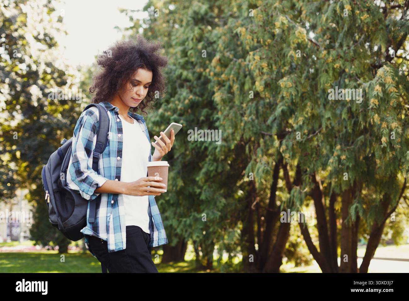 I giovani si godono un caffè mentre controllano il telefono in un parco durante il giorno con gli alberi verdi Foto Stock