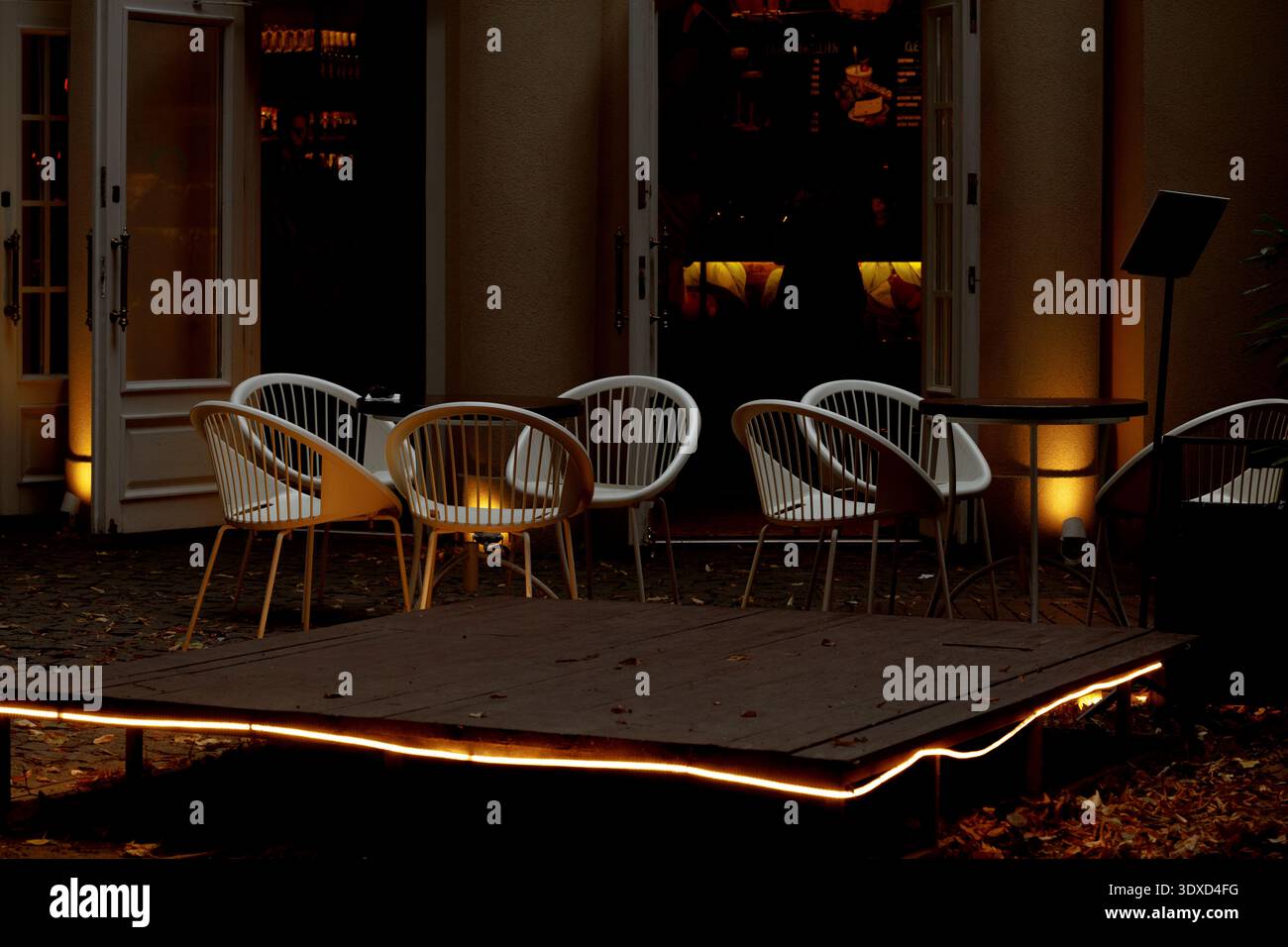 Sedie e tavoli bianchi vuoti sono disposti su un patio, inondati dal soffice bagliore dell'illuminazione ambientale mentre il crepuscolo si deposita Foto Stock