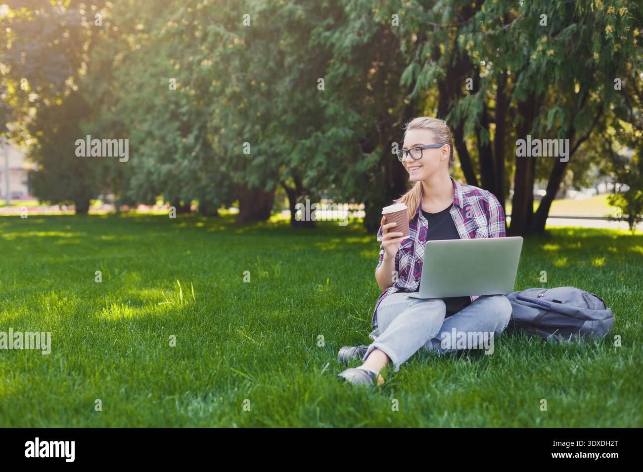 Donna che usa un laptop e gusta un caffè in un parco verde durante il giorno Foto Stock