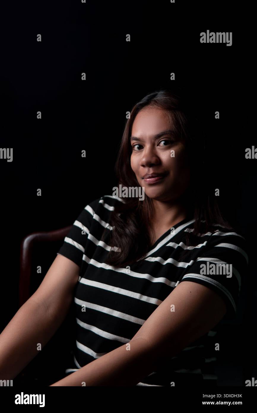 Una donna indonesiana sicura con una camicia a righe che posa con un sorriso sottile. Ritratto scuro cinematografico per il branding personale e i concetti di narrazione. Foto Stock