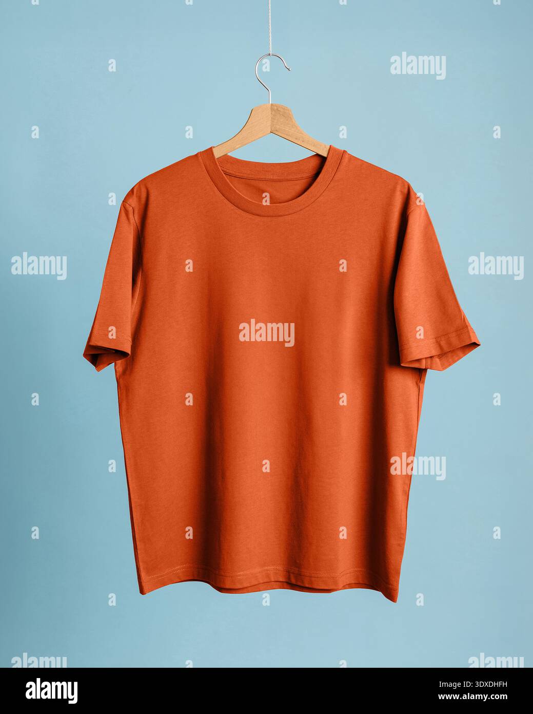 T-shirt in cotone arancione bruciato appesa su un gancio di legno su sfondo blu polvere. La t-shirt è semplice, a maniche corte e leggermente morbida Foto Stock