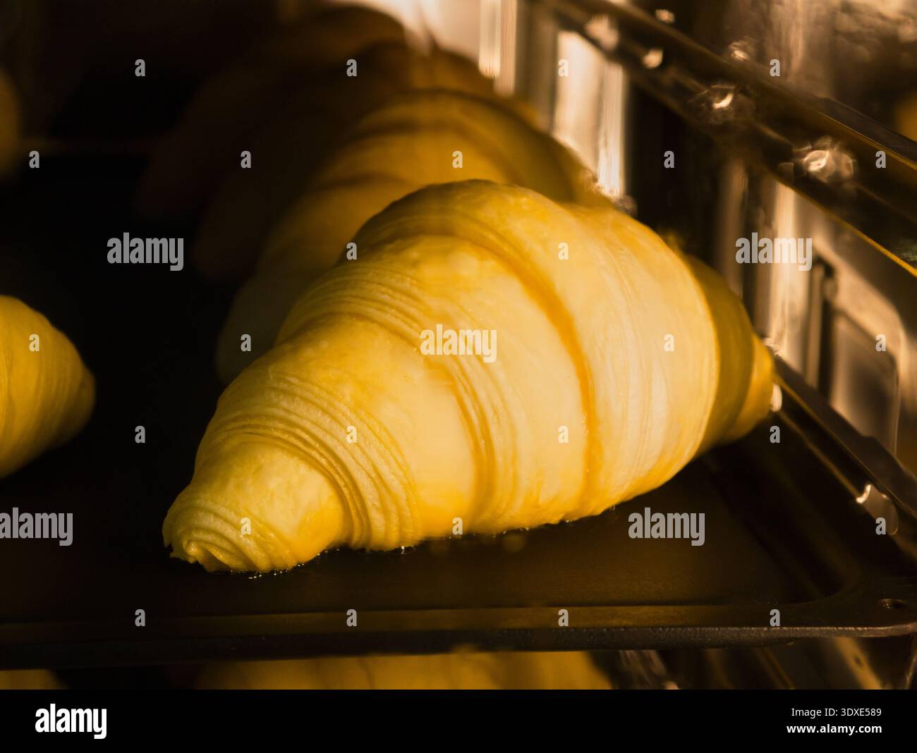 I croissant dorati si alzano e cuocono alla perfezione in un vivace forno commerciale durante l'ora di punta mattutina per le prelibatezze della colazione Foto Stock