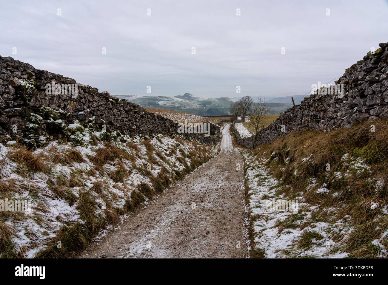 Il sentiero di ghiaia ricoperto di gelo scende tra alti muretti a secco verso un lontano fienile di pietra e colline innevate nelle Dales dello Yorkshire Foto Stock
