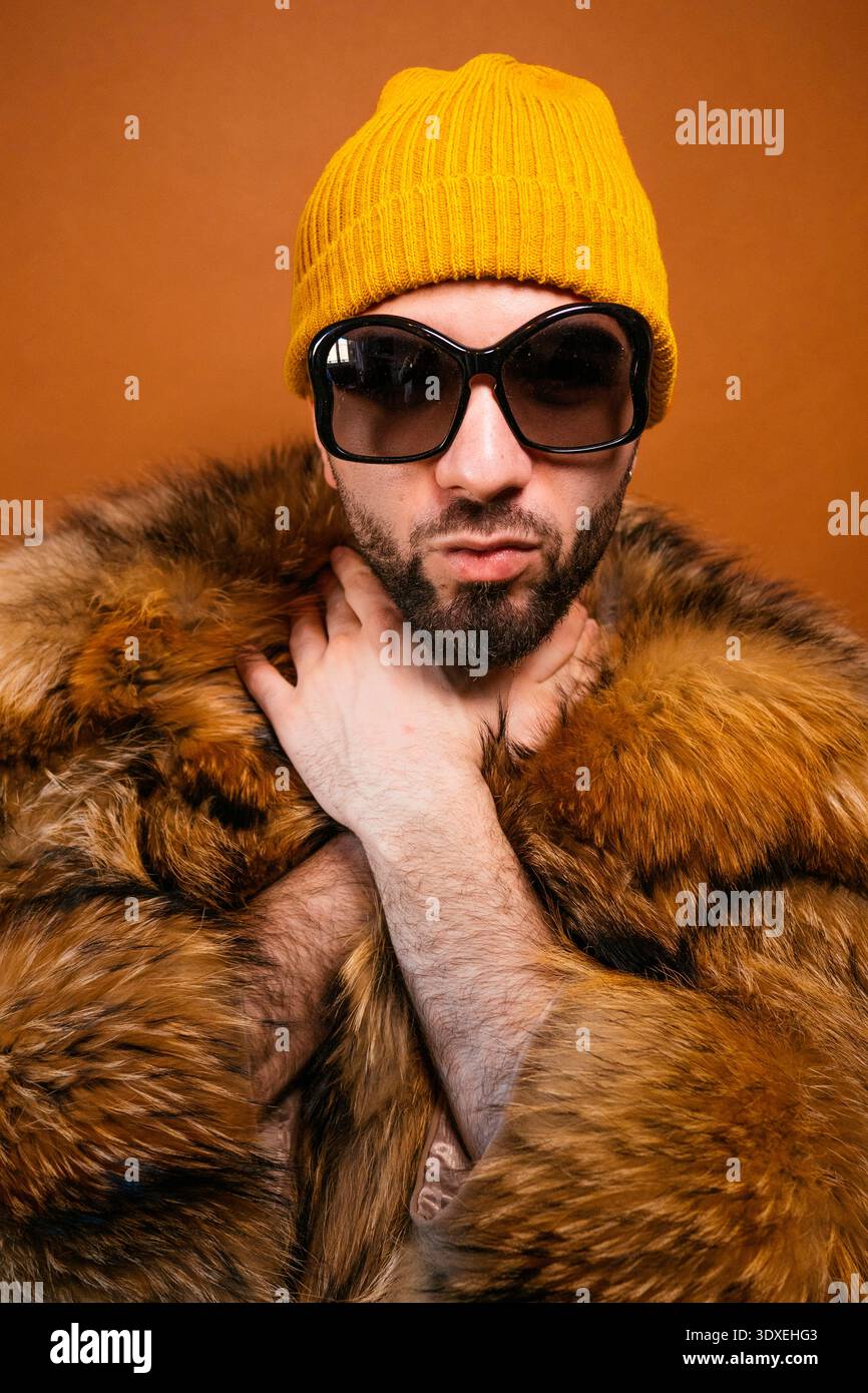Giovane adulto con un cappotto di pelliccia, un berretto giallo e occhiali da sole su sfondo marrone Foto Stock