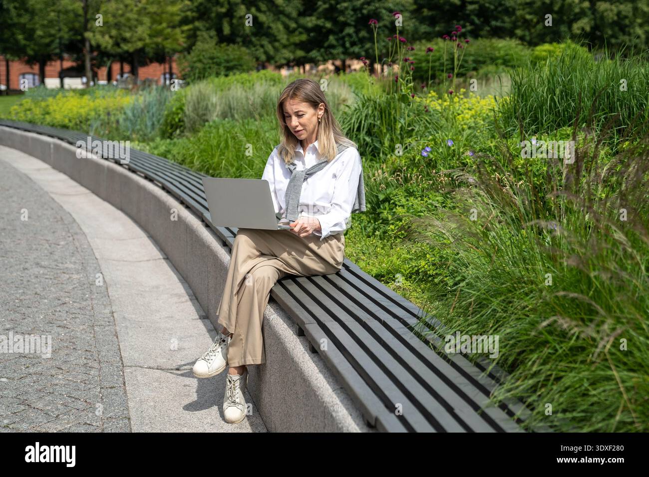 Donna d'affari positiva che lavora al telecomando in un parco, seduto sulla panchina che parla con una videochiamata sul laptop. Foto Stock