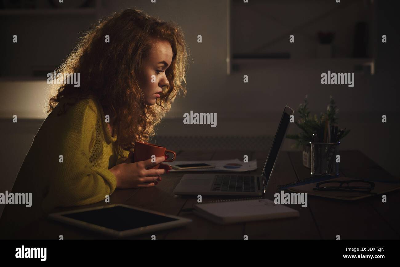 Donna che fissa il laptop con un caffè in mano durante le ore serali a Home Space Foto Stock