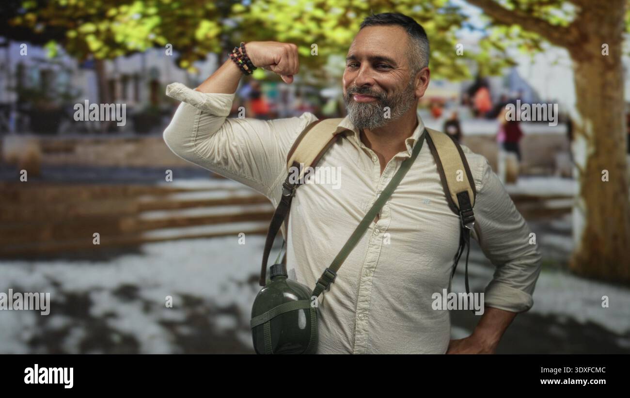 L'uomo ispanico di mezza età si flette bicipite, sorridendo, indossando zaino con mensa e braccialetti in Street plaza; avventura sicura. Foto Stock