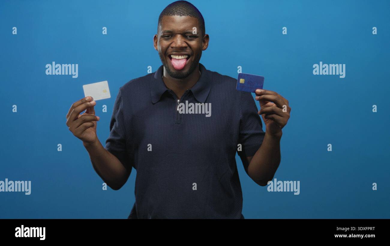 Giovane con due carte di credito, linguetta a occhi chiusi che le presenta sullo sfondo blu dello studio; scelta giocosa. Foto Stock
