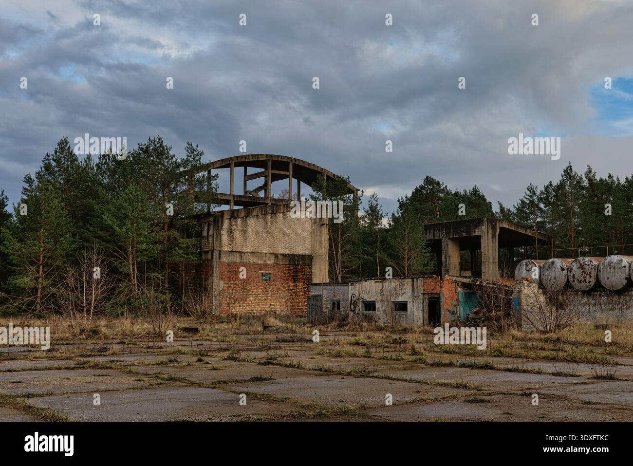 Immagine ampia di un sito industriale abbandonato o di un complesso militare in una foresta, con strutture in cemento in decadenza e muri in mattoni sotto un cielo nuvoloso. Foto Stock