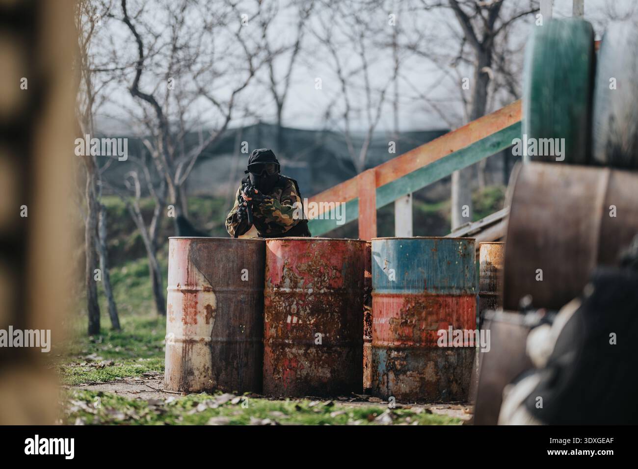 Il giocatore di paintball accovacciato dietro barili arrugginiti, puntando un marcatore in un campo all'aperto Foto Stock