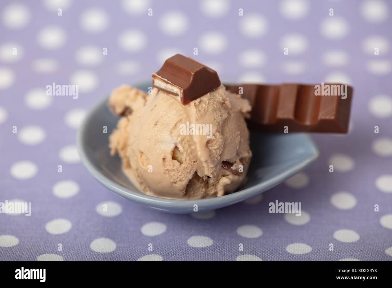 Una paletta di gelato al cioccolato leggero con pezzi di cioccolato al latte visibili, servita in una piccola ciotola blu su uno sfondo a pois Foto Stock