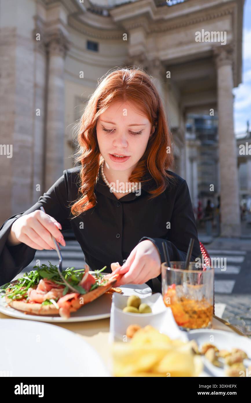 Elegante donna rossa che gusta un aperitivo al caffè all'aperto di Roma. Uno stile di vita da viaggio soleggiato e un'atmosfera da vacanza italiana. Foto Stock