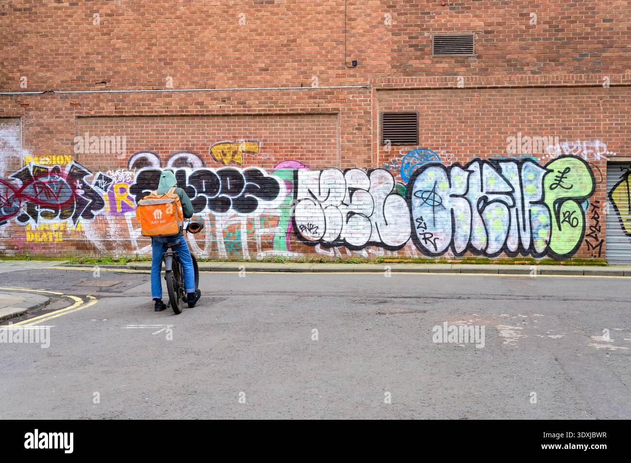 Un ciclista che consegna il cibo appena mangiato stazionario su una strada della città con graffiti su un muro, nel Regno Unito Foto Stock