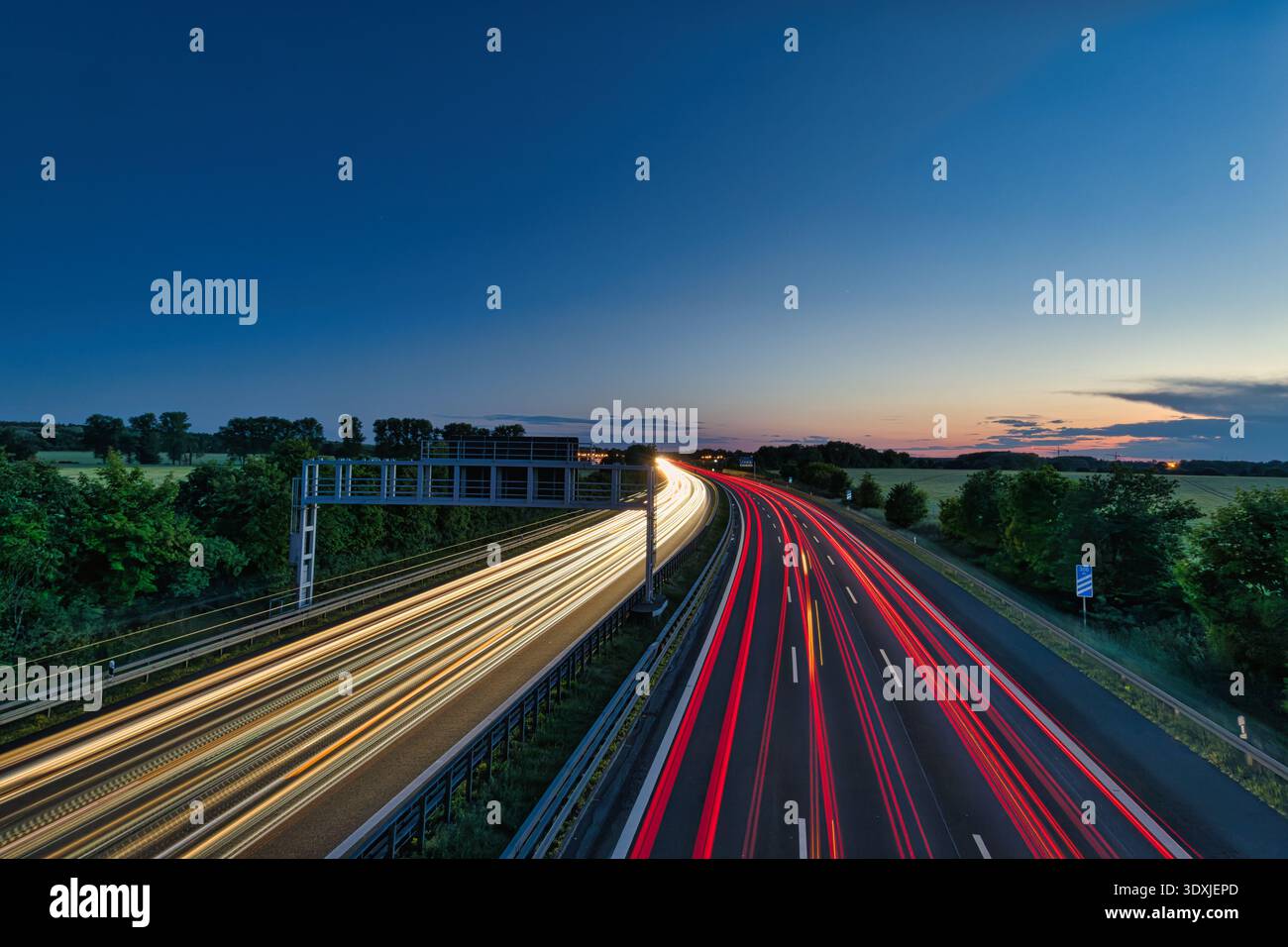 Lunga esposizione del traffico autostradale al tramonto, con percorsi leggeri dai fari e dalle luci posteriori dell'auto contro il cielo del tramonto e le sagome degli alberi Foto Stock