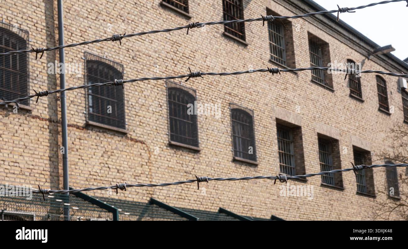 Vecchio edificio carcerario con sbarre sulle finestre e recinzione di filo spinato in primo piano. Foto Stock