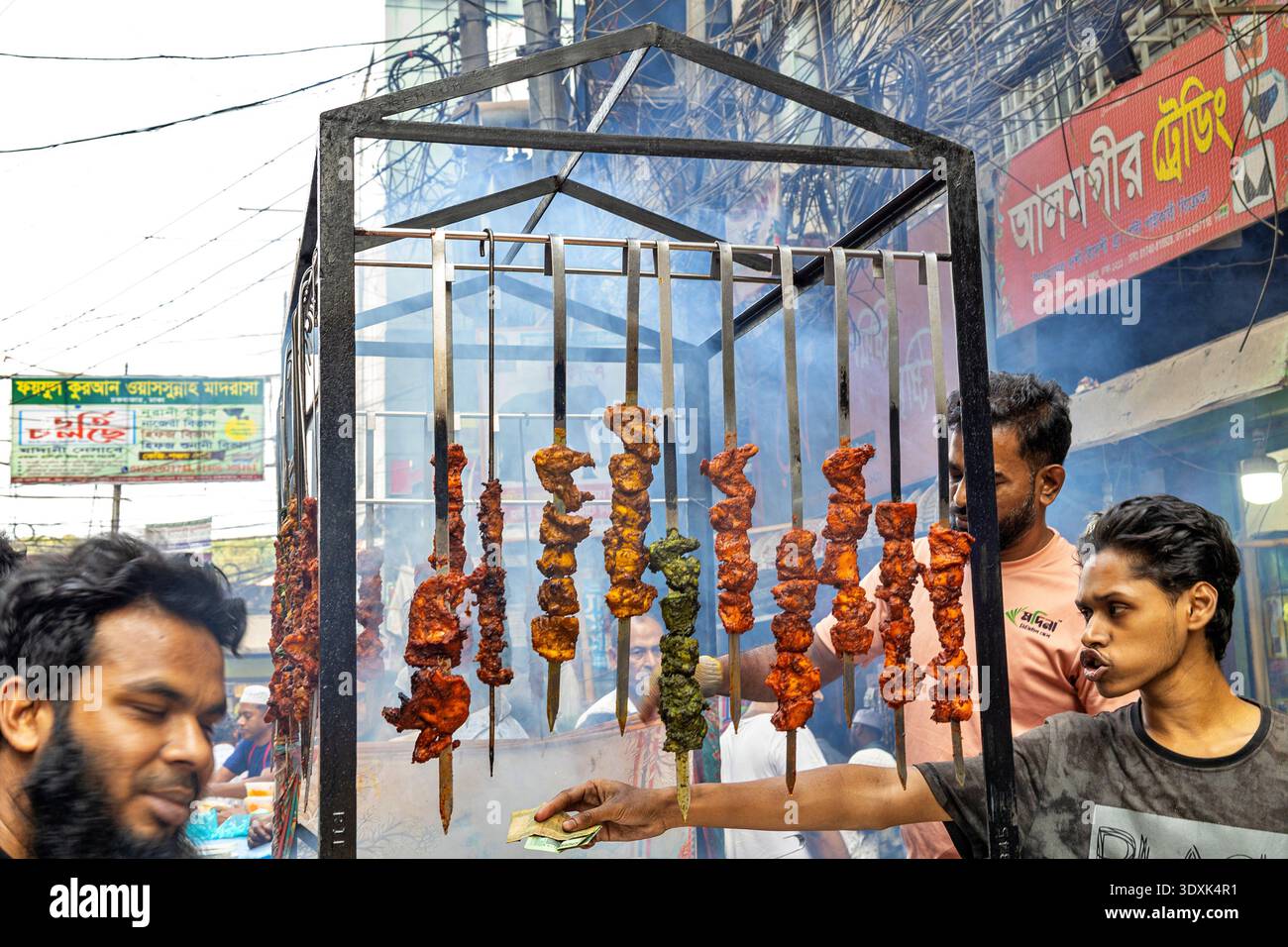 Vista di spiedini sfrigolanti appesi a una struttura in metallo, fumo che sale intorno ai venditori e ai clienti nella vivace strada di Chawkbazar, Dhaka, Bangladesh. Foto Stock