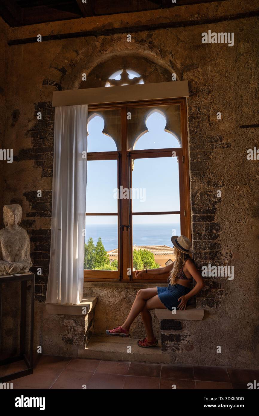 Donna seduta accanto a una finestra ad arco che guarda al mare in interni storici Foto Stock
