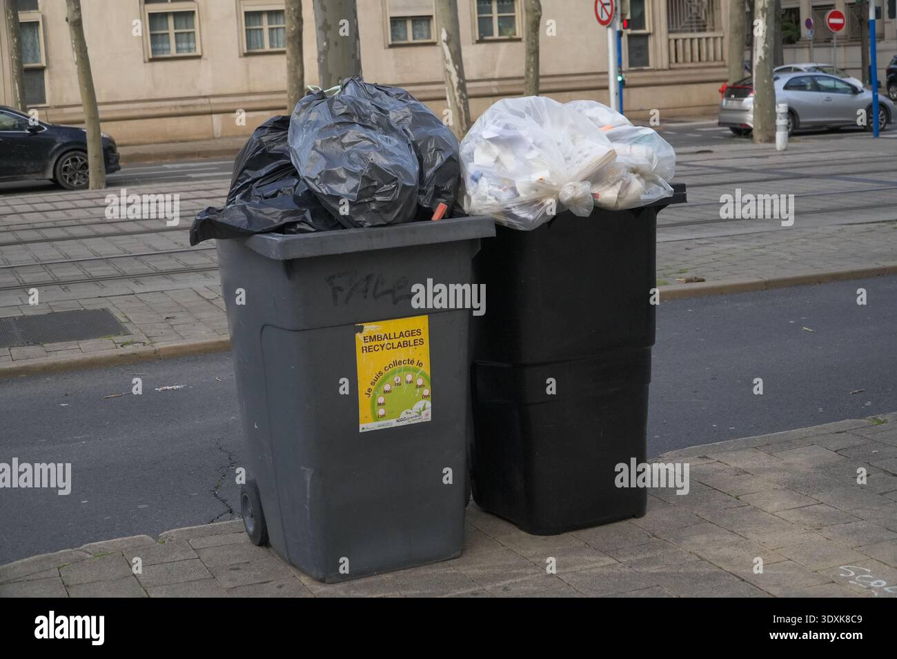 I contenitori pieni di rifiuti, compreso un cestino di riciclo grigio con etichetta francese, si trovano sul marciapiede a Montpellier. Concetto di gestione dei rifiuti. Foto Stock