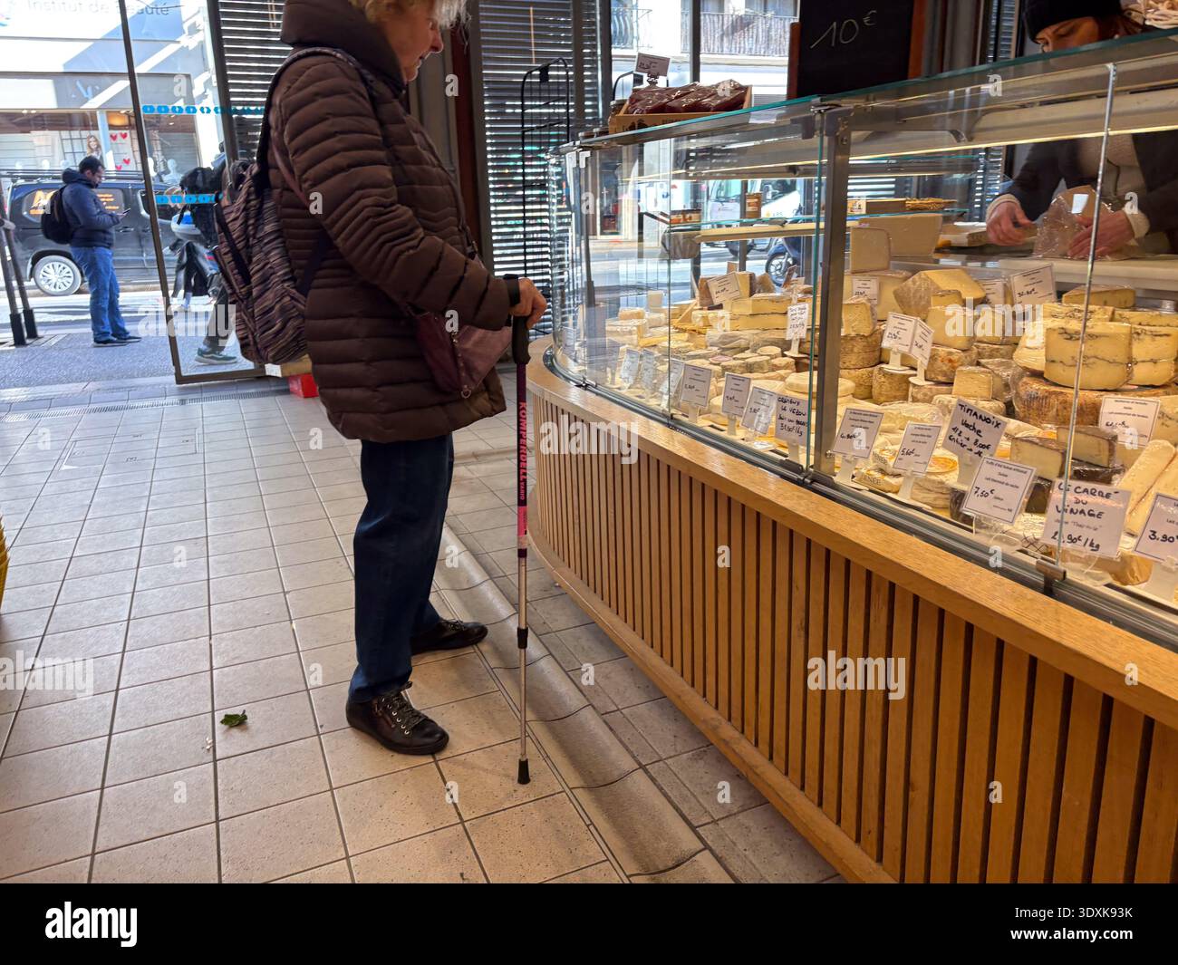 Donna anziana che fa shopping usando una canna pieghevole mentre sfoglia formaggi artigianali in una fromagerie francese. Scena di vita quotidiana. Foto Stock